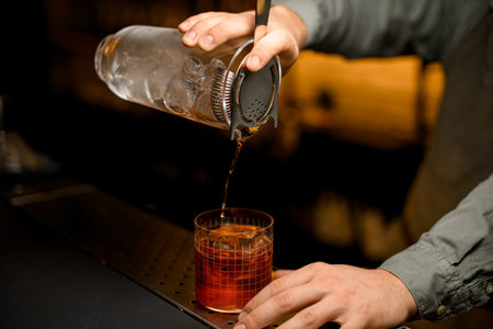 hand of bartender neatly holds mixing cup with strainer while pouring drink into glass