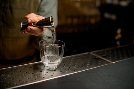 hand of bartender holds jigger and pours drink into transparent glass with iceの写真素材