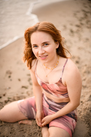 portrait of caucasian smiling beautiful red-haired young woman sitting on sand of beach.の写真素材