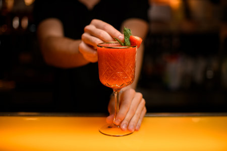 close-up of goblet with signature cocktail which hand of bartender decorate with mint leaf and...