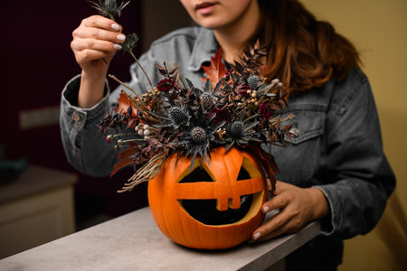 female hand gently decorate arrangement of thistles flowers and plants in pumpkin with carved eyes and mouth. Decor for halloween.の写真素材