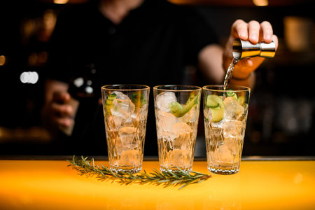 hand of bartender carefully pours drink from steel jigger into glass with ice