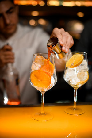 Close-up of glass with ice cubes and orange slices on the bar and bartender pours alcoholic drink from jigger into glassの写真素材