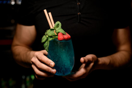 hands of man bartender accurate holds glass with blue cocktail decorated with mint and red raspberries. Cropped photoの写真素材