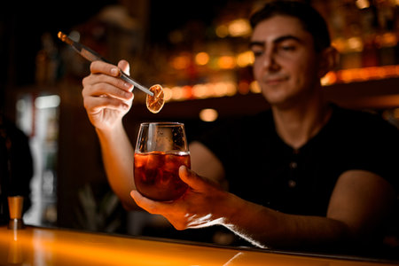 male bartender gently holds slice of dry citrus with tweezers over glass with cold cocktailの写真素材