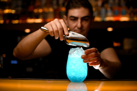magnificent view of glass with blue cocktail on bar and metal scoop in hands of bartender above itの写真素材