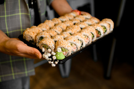 Tasty sushi set of california rolls with salmon garnished sesame seeds in black tray in male hands.の写真素材