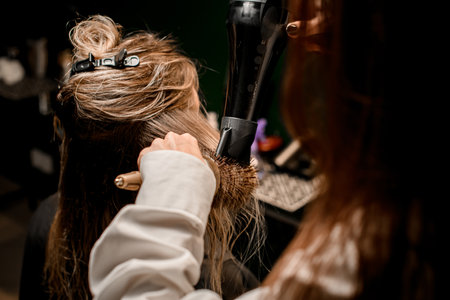 Female hairdresser stylist skilfully making hairstyle using hair dryer and round combの写真素材