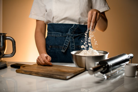 hand of woman pouring white flour in stainless bowl on table with modern kitchen appliancesの写真素材