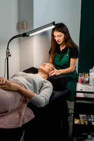 professional woman beautician makes face massage for female client in beauty salon.