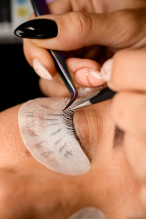 close-up of beautician gently attaches artificial eyelashes with tweezers. Eyelash extension...
