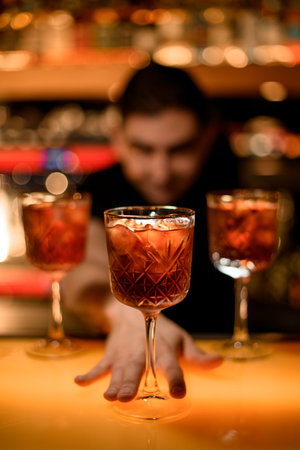 Male bartender presents cold red cocktail with ice cubes in glass on the bar counter in the barの写真素材
