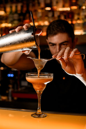 barman holds sieve in his hand over champagne glass and gently pours drink from steel shaker cup.の写真素材
