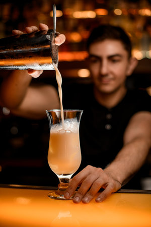 barman holds sieve in his hand over cocktail glass and gently pours drink from steel shaker cup.の写真素材
