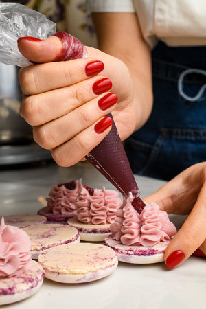 Chef woman squeezing fruit jam into ganache cream on macarons halves, close up