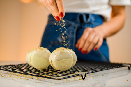 Woman confectioner with bright red manicure sprinkles freshly made macaroons with chopped peanuts and pistachiosの写真素材