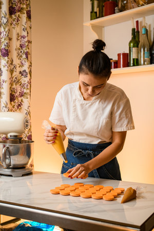 Woman with confectionery bag squeezing cream filling to macarons shells at kitchen interiorの写真素材