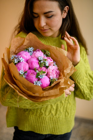 Young beautiful woman in green sweater posing with bouquet of peonies and blue small decorative flowers in stairwell interiorの写真素材