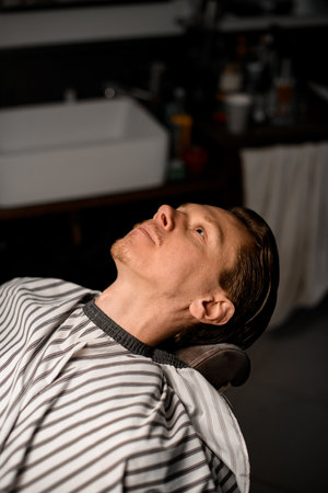 Handsome young bearded guy with heardressers cape on sitting in armchair in barber shopの写真素材
