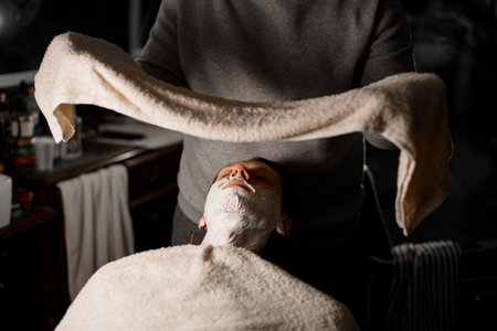 Hands of barber applying hot towel to male clients face to moisturize beard hairs before shavingの写真素材