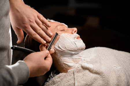 Male barber holding vintage straight razor in hands and shaving young attractive clientの写真素材