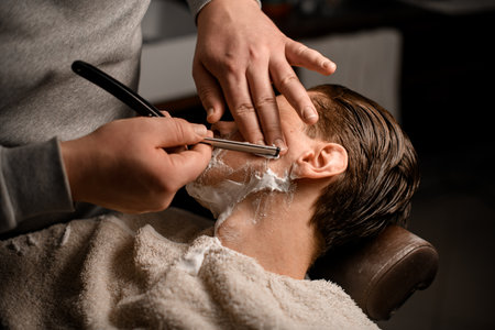 Barber man shaving young attractive client using vintage straight razor with stainless steel in his salonの写真素材