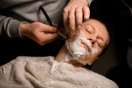 Young attractive man relaxing while sitting in barbers chair and getting shave by male barberの写真素材