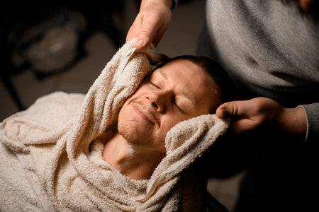 Barber covering clients face with hot towel before giving him shave at barbershop.の写真素材