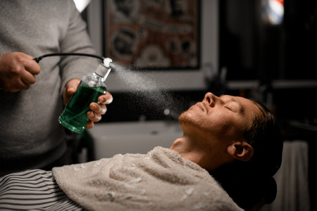Man hairdresser sprinkles perfume on clients face in modern barbershop on black background.の写真素材