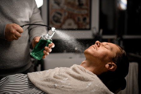 Barber sprinkles perfume on clients face in modern barbershop on blurred background.の写真素材