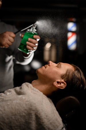 Man barber sprinkles perfume from vintage desidned stylish bottle above clients face in modern barbershopの写真素材