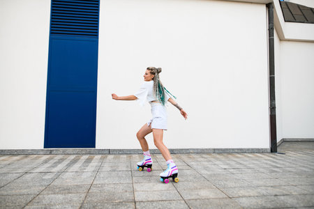 Young woman with colorful braids in hair skates on retro rollers outside against white wall...