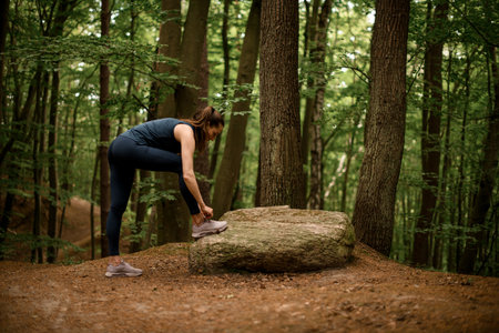 Woman runner stands near large stone and tying her shoelaces, ready to run through forestの写真素材