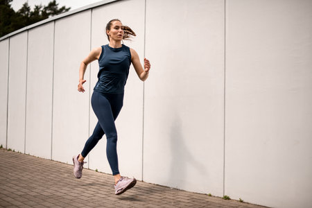 Front view of sporty young woman running on sidewalk in morning. White wall background
