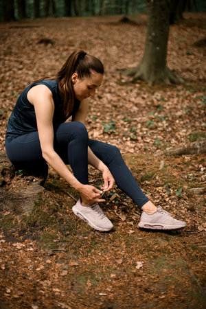 Woman runner sits on tree stump and tying her shoelaces, ready to run through forestの写真素材