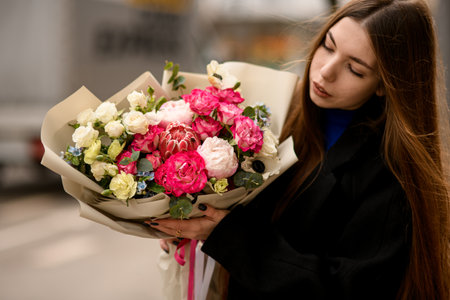 Young woman in black coat holding large luxurious bouquet of flowers with protea, roses, peonies wrapped in craft paperの写真素材