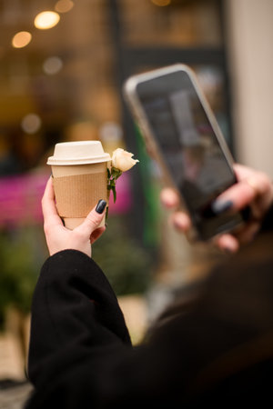 Female hand taking photo of paper cup with takeaway coffee decorated with rose flower on smartphone,...