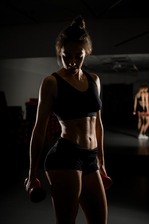 Adorable fitness woman with lifting dumbbells showing her well trained body, black backgroundの写真素材