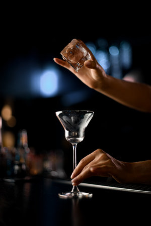 Unrecognizable bartender putting big ice cube into glass while preparing cocktail in bar. Blurred background. Closeup viewの写真素材
