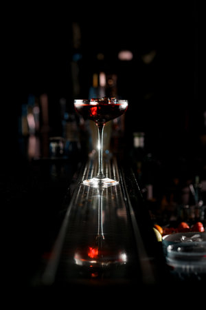 View on tasty red cocktail in high martini glass with ice cube reflected in steel surface of bar counter. Dark blurred background. Menu of classic cocktailsの写真素材
