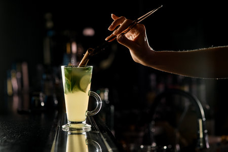 close-up on cocktail glass with citrus liquid and woman's hand holding tweeters and pouring stick of cinnamon into it. Dark background. Front viewの写真素材