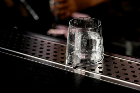 Beautiful view of transparent old-fashioned glass with ice cube standing on bar counter. Bartender's silhouette on blurred backgroundの写真素材