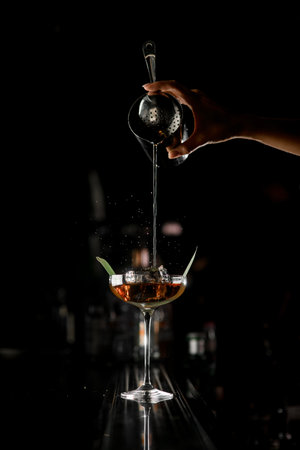 View on bar counter with transparent crystal glass in which female bartender's hand pours cocktail from steel mixing cup. Concept about professions and drinksの写真素材