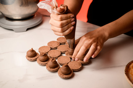Female hands of a confectioner squeeze brown cream onto beige halves of macaroons