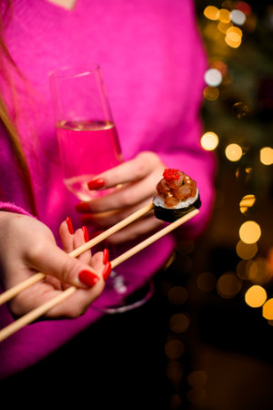 Female hands hold a sushi roll with chopsticks and a glass of champagne against a background of glowing lightsの写真素材
