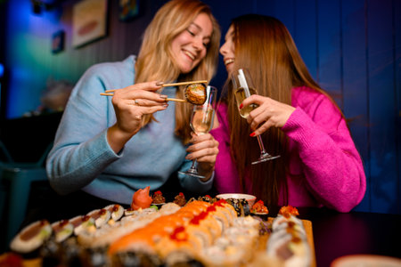 Two cute happy women happily eating sushi rolls with chopsticks and holding glasses of champagneの写真素材