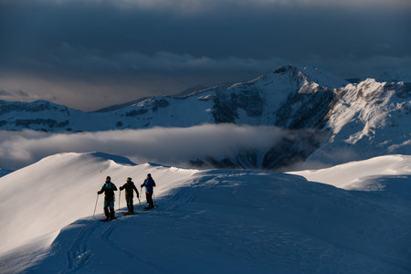 Three male skiers stand on a snowy peak of a mountain, only part of which is illuminated by the suns...