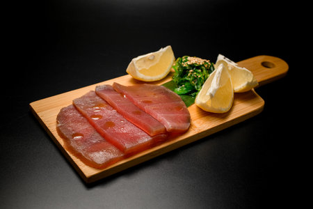 Flat slices of tuna fillet, lemon wedges and seaweed with sesame lie on a wooden boardの写真素材
