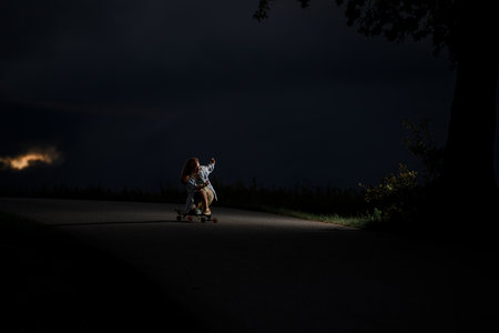 Young girl is riding along the road crouching on a longboard next to a single tree with light coming from itの写真素材