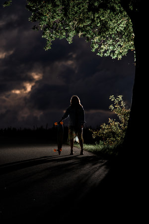 Silhouette of a young girl with a longboard standing in the dark on the road, behind her is illuminated by additional lightの写真素材